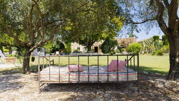 Una cama con estructura metálica y colchón a rayas, rodeada por una zona de grava y árboles.