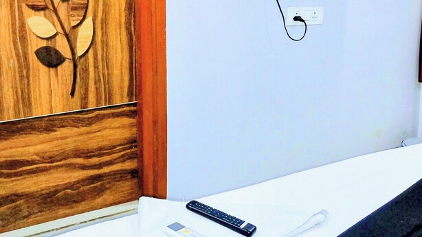 A room with a wooden door, a flat-screen TV mounted on the wall, a white desk, and a remote control on the desk. - THE ROYAL STAY IN (Hyderabad)