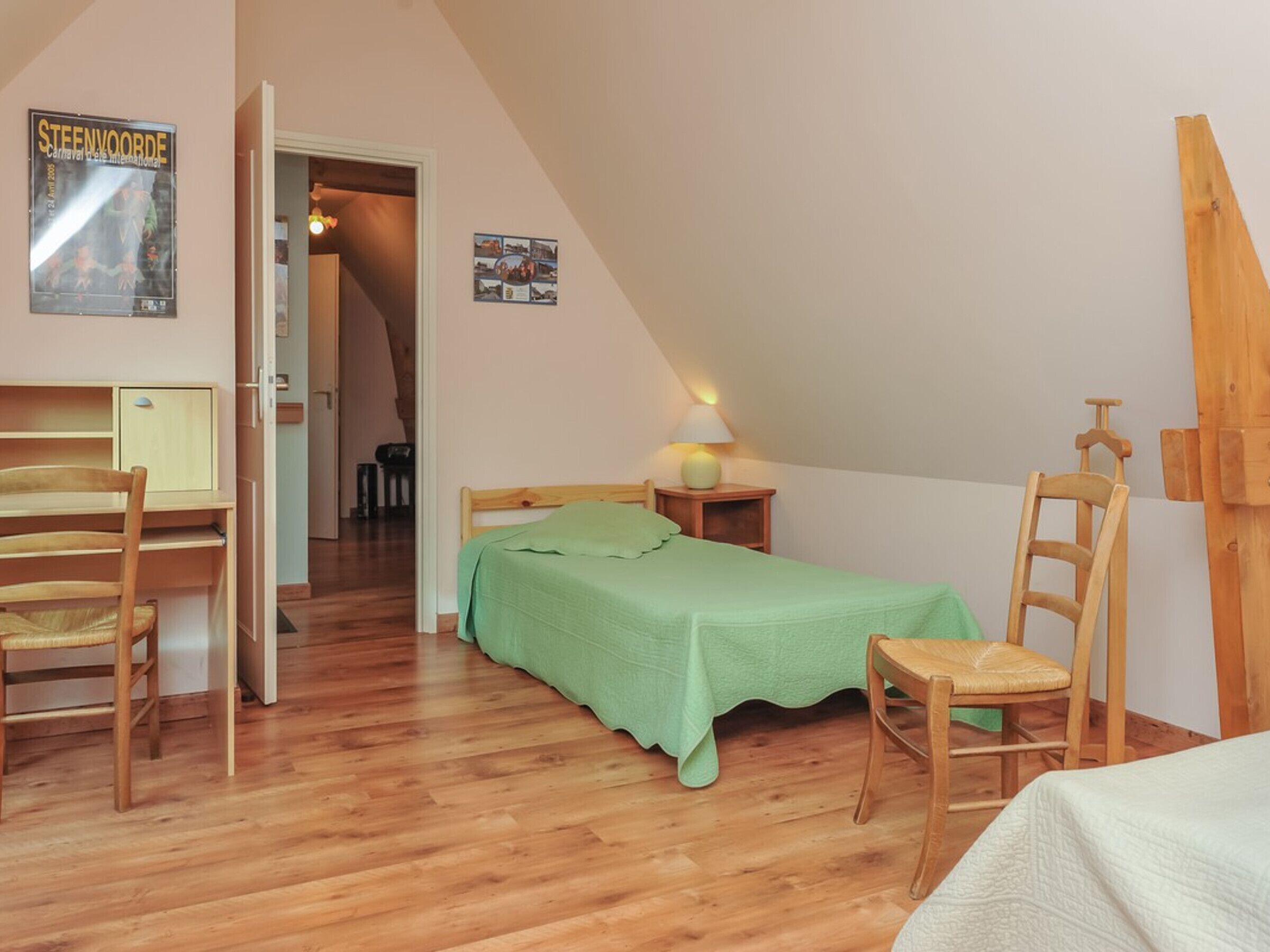A cozy, well-lit room with a wooden bed, a desk, and a chair. A poster is visible on the wall, and a ladder is leaning against the wall.