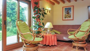 A room with wicker chairs, a small table with a lamp, and a view of greenery outside.