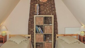A cozy attic bedroom with two single beds, a wooden ceiling, a brick wall, and a bookshelf.