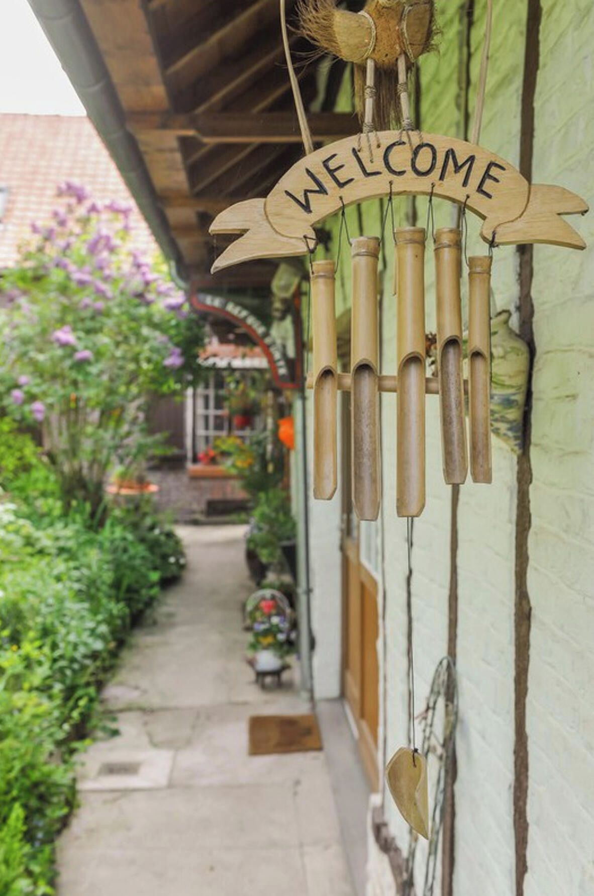 Ein hölzernes Schild mit dem Wort 'WELCOME', das an einer Konstruktion, möglicherweise einem Eingangsbereich oder einer Veranda, hängt, mit einem Gartenweg, der zu einem Gebäude im Hintergrund führt.