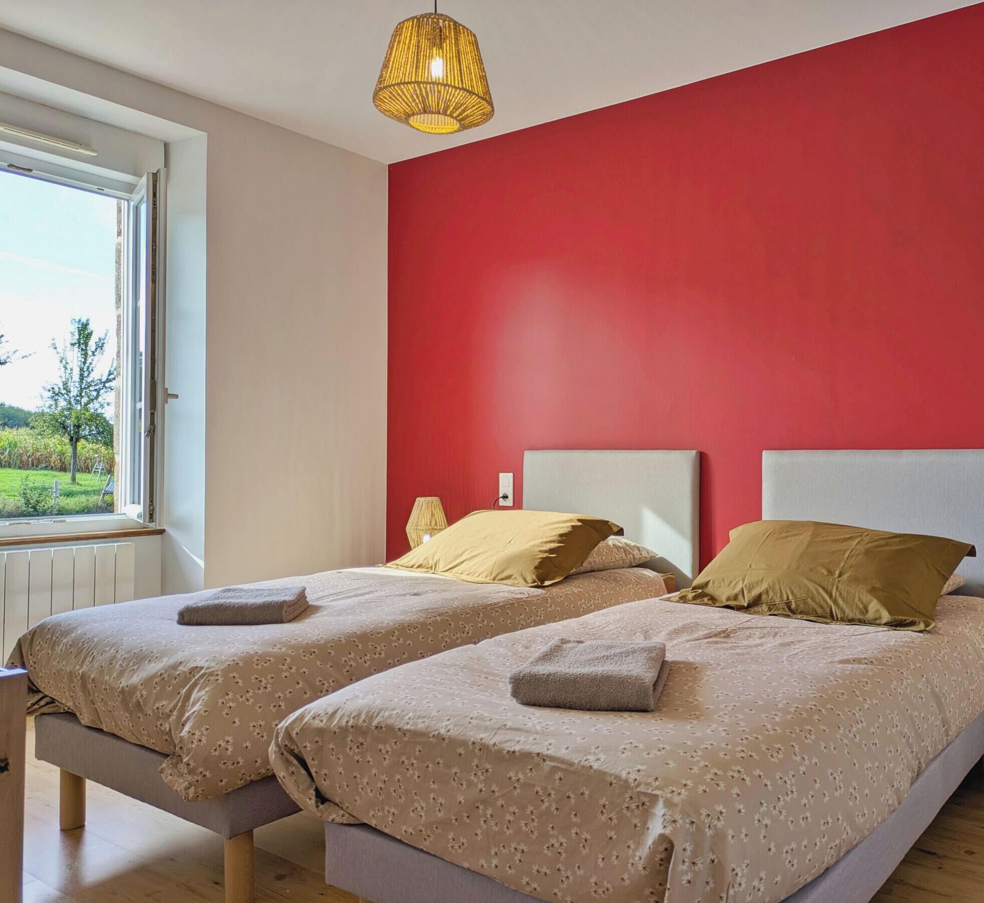 A room with two beds, a red wall, and a window with a view of greenery.