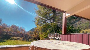 Ein Tisch mit gemustertem Tischläufer und drei Weingläsern, vor einer malerischen Kulisse aus Bäumen und Bergen.