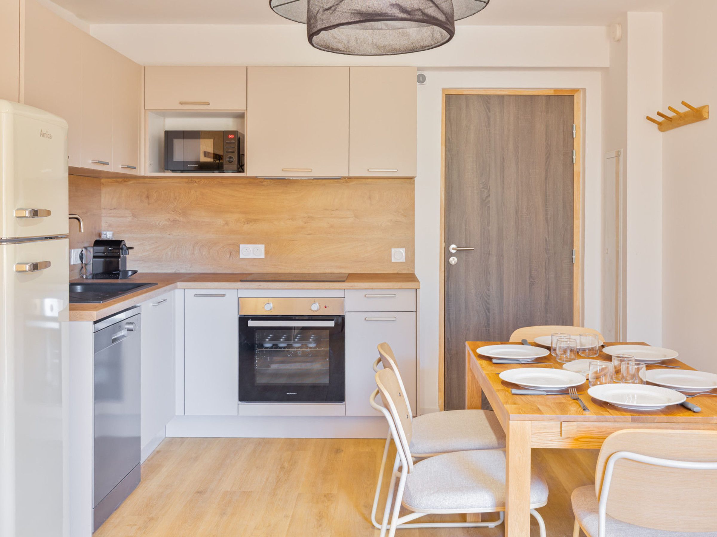 Une cuisine compacte dotée d’appareils électroménagers modernes, un coin repas avec une table et des chaises en bois, et une porte donnant sur une autre pièce.
