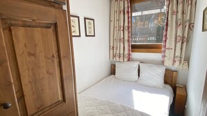 A bedroom with a wooden door, a bed with white bedding, a window with floral curtains, and framed pictures on the wall.
