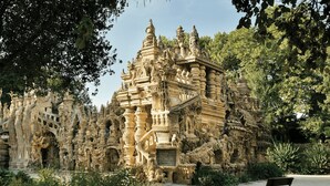 A detailed stone structure with intricate carvings, surrounded by greenery and a bench in the foreground. - Studio in the heart of the village (Hauterives)