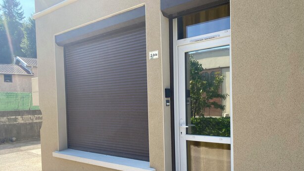 A building with a closed roller shutter door and a window with a view of greenery. - Studio in the heart of the village (Hauterives)