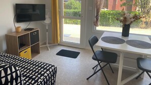 A modern room with a TV, a desk with two chairs, a sofa, a fan, and a window with a view of greenery and buildings. - Studio in the heart of the village (Hauterives)
