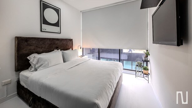 Une chambre moderne avec un grand lit, une tête de lit, un téléviseur fixé au mur et un balcon agrémenté de plantes.