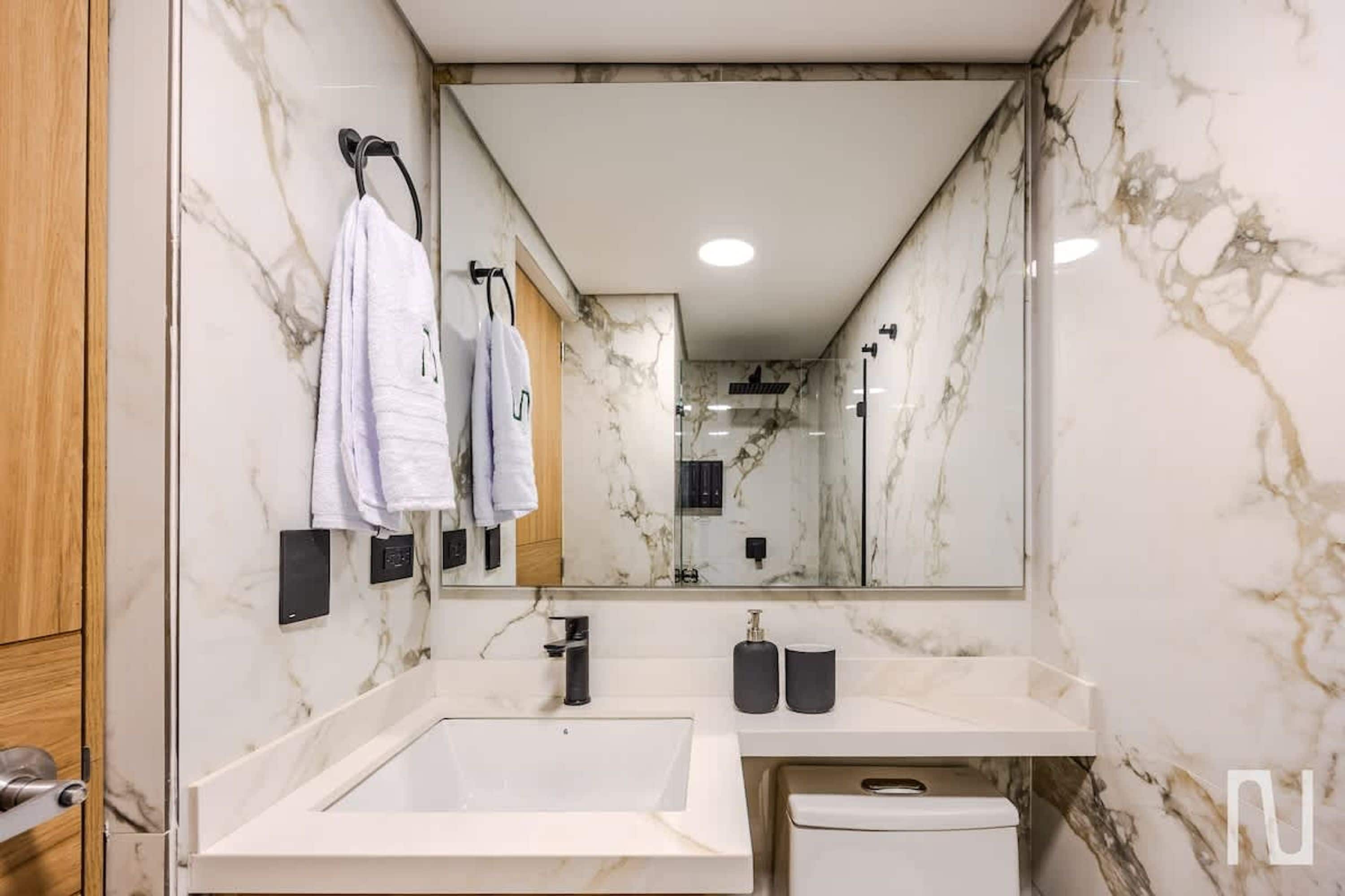 Ein modernes Badezimmer mit einem großen Spiegel, Marmorwänden, einem weißen Waschbecken und einer Toilette.