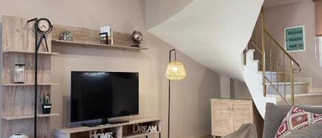 A modern living room with a flat-screen TV, a coffee table, and a sofa. There is a staircase leading to an upper level.