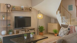 A modern living room with a flat-screen TV, a coffee table, and a sofa. There are decorative items on shelves and a staircase in the background.