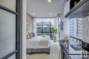 Habitación de hotel moderna con una cama grande, una pequeña kitchenette, un ventanal amplio con vistas a la ciudad y una planta en maceta.