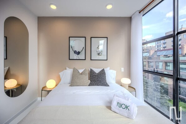 Habitación de hotel con una cama grande, dos mesitas de noche con lámparas, un ventanal con vistas a la ciudad y cuadros en la pared.