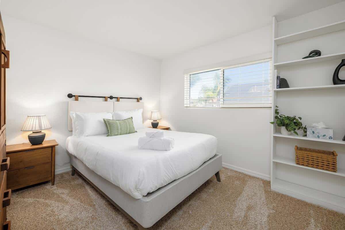 A neatly arranged bedroom with a bed, bedside tables, a window, and a built-in shelf with various items.