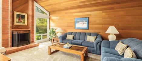 A living room with a fireplace, blue sofa, wooden coffee table, and a large window.