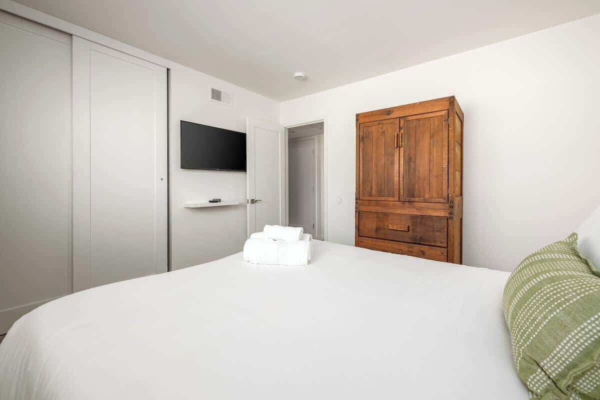 Ein Schlafzimmer mit einem Bett, einem Holzschrank, einem an der Wand montierten Fernseher und einer Tür zu einem anderen Raum.