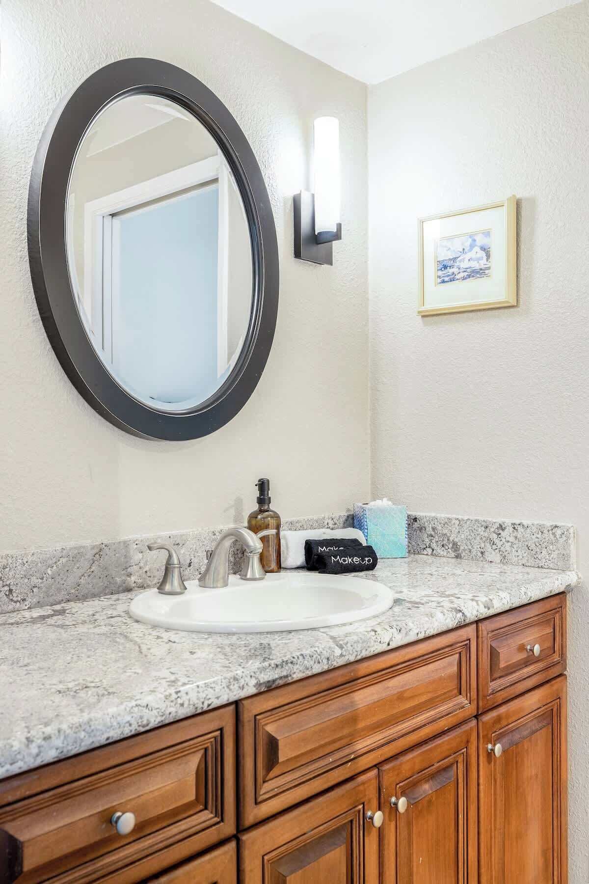 A bathroom with a round mirror, a sink, wooden cabinets, a towel, and a framed picture on the wall.