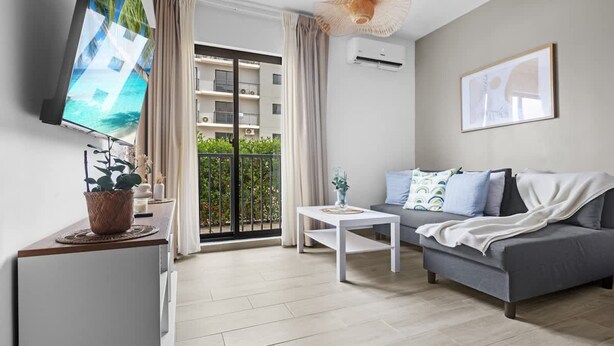Ein modernes Wohnzimmer mit grauem Sofa, einem weißen Couchtisch, einem Flachbildfernseher und einem Balkon mit Blick auf ein Gebäude und Grünflächen.