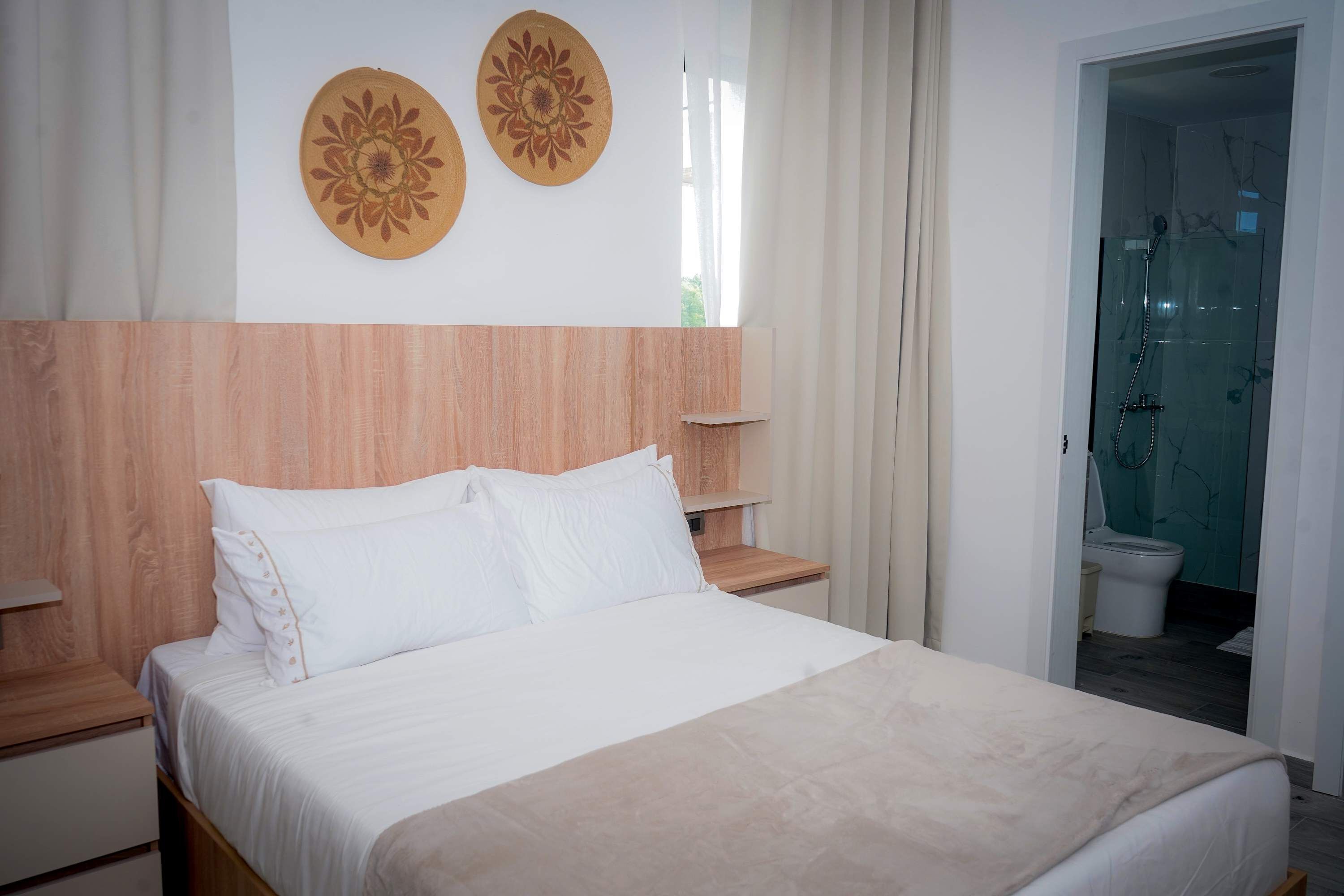 Hotellihuone, jossa on puinen sängynpääty, valkoisilla lakanoiden koristeltu sänky, kaksi seinälle kiinnitettyä koristeellista lautasparia, puinen yöpöytä ja lasiseinällä erotettu suihkutila.
