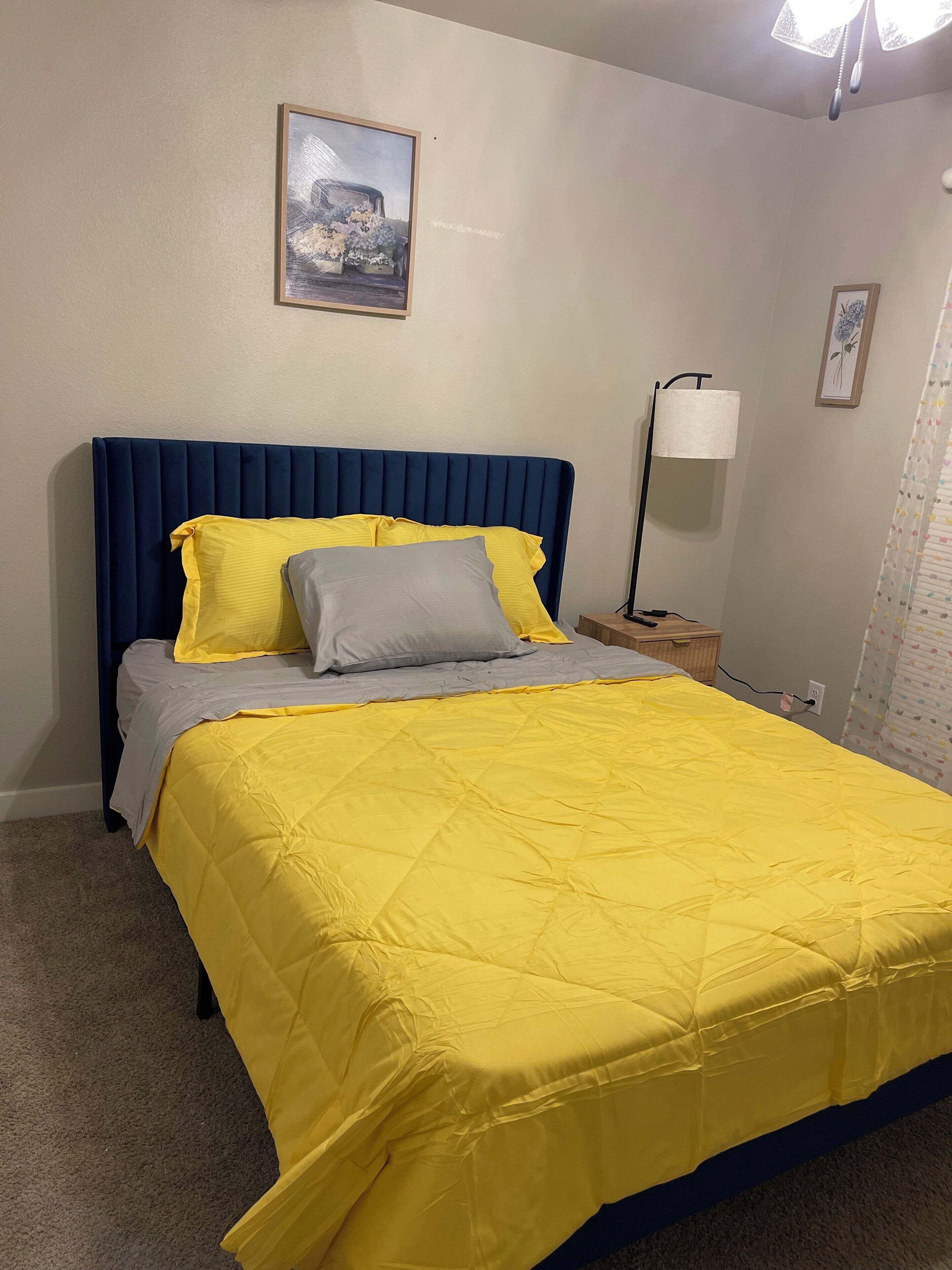 Una cama con cabecera azul, ropa de cama amarilla y gris, una lámpara de noche y un cuadro en la pared.