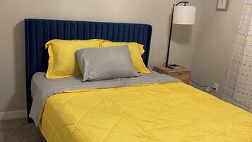 Una cama con cabecera azul, ropa de cama amarilla y gris, una lámpara de noche y un cuadro en la pared.