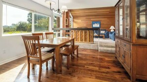 Een ruime eethoek met een houten tafel en stoelen, een keuken met houten kastjes, en uitzicht op de omgeving door grote ramen.