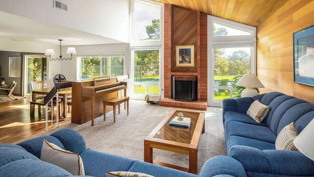 Een ruime woonkamer met een piano, een open haard en grote ramen met uitzicht op de omgeving.