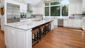 Una cocina amplia con armarios blancos, una isla grande con taburetes, un refrigerador de acero inoxidable y una ventana con vista a árboles.
