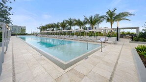 En swimmingpool med solstolar och parasoller, omgiven av palmer och moderna byggnader.