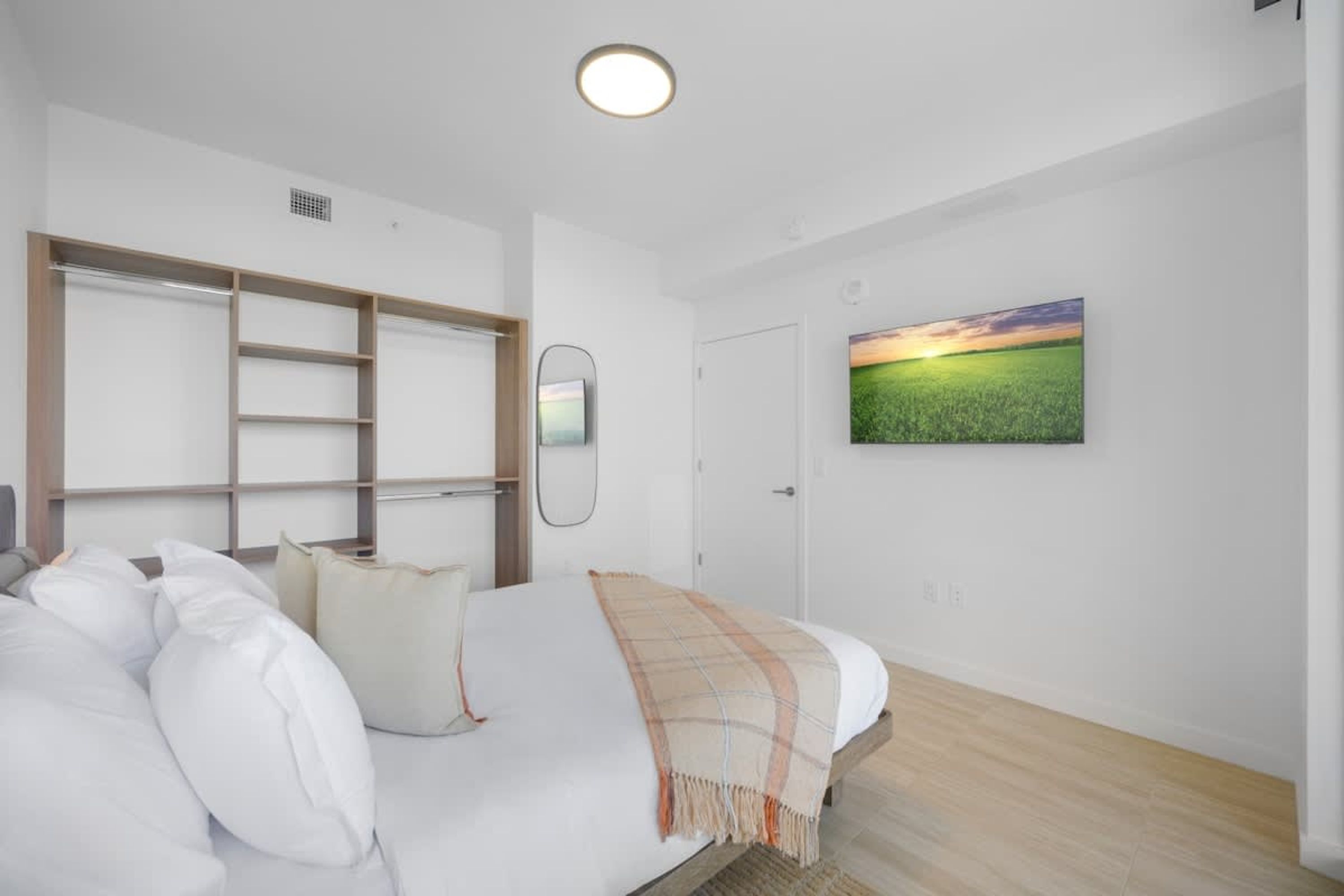 A bedroom with a bed, a wardrobe, a mirror, and a painting of a field.