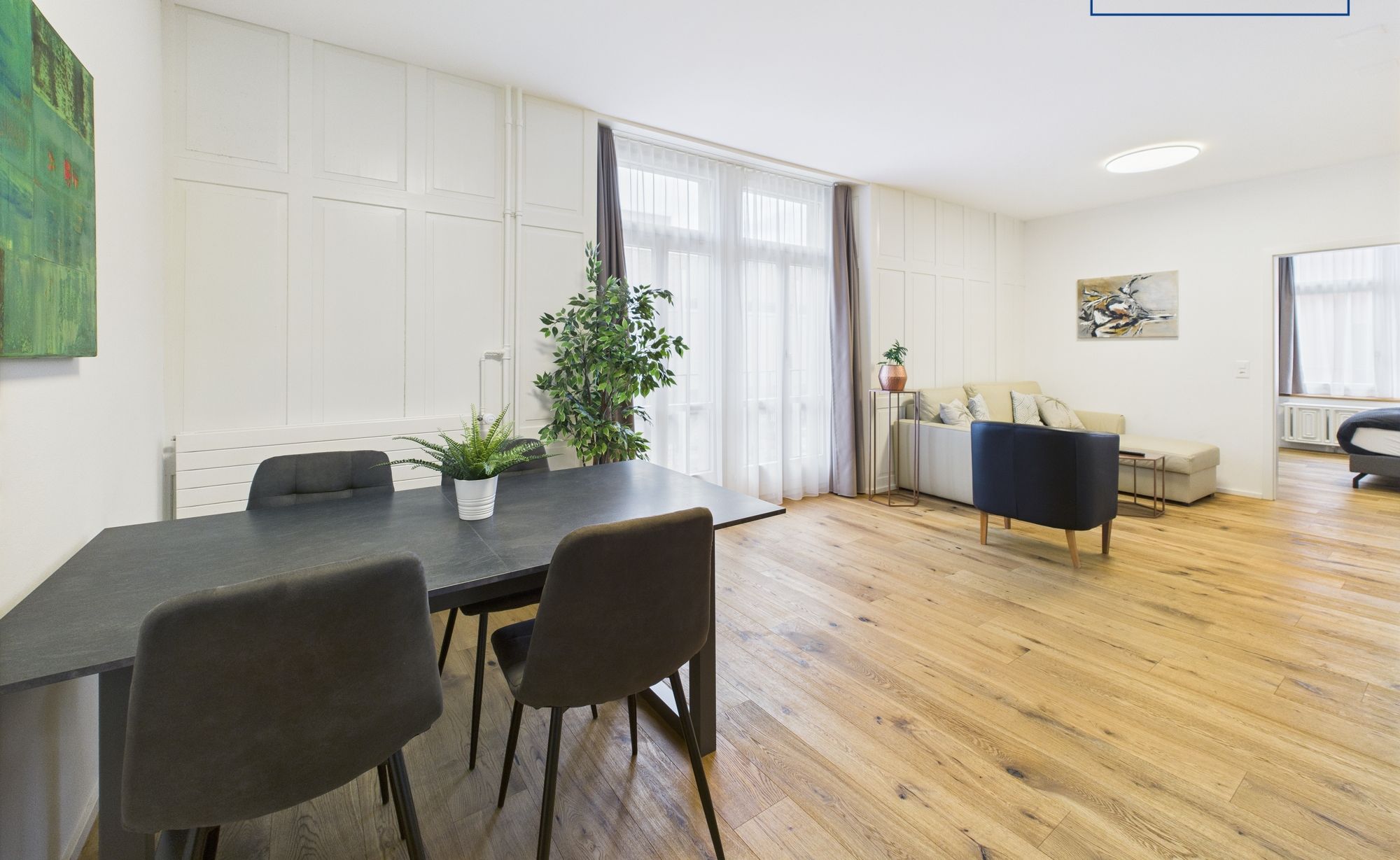 A modern living room with a dining area, wooden flooring, and a large window with curtains.