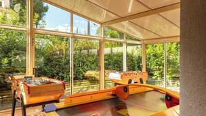 A sunlit room with a foosball table and a pool table, large windows offering a view of greenery outside. - Camping Marvilla Parks - Clos Cottet (Angles)