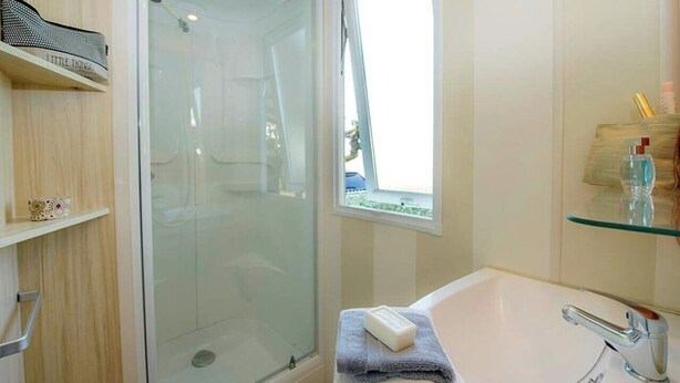 A bathroom with a glass shower enclosure, a white sink with a chrome faucet, and a window providing natural light. - Camping Marvilla Parks - Clos Cottet (Angles)