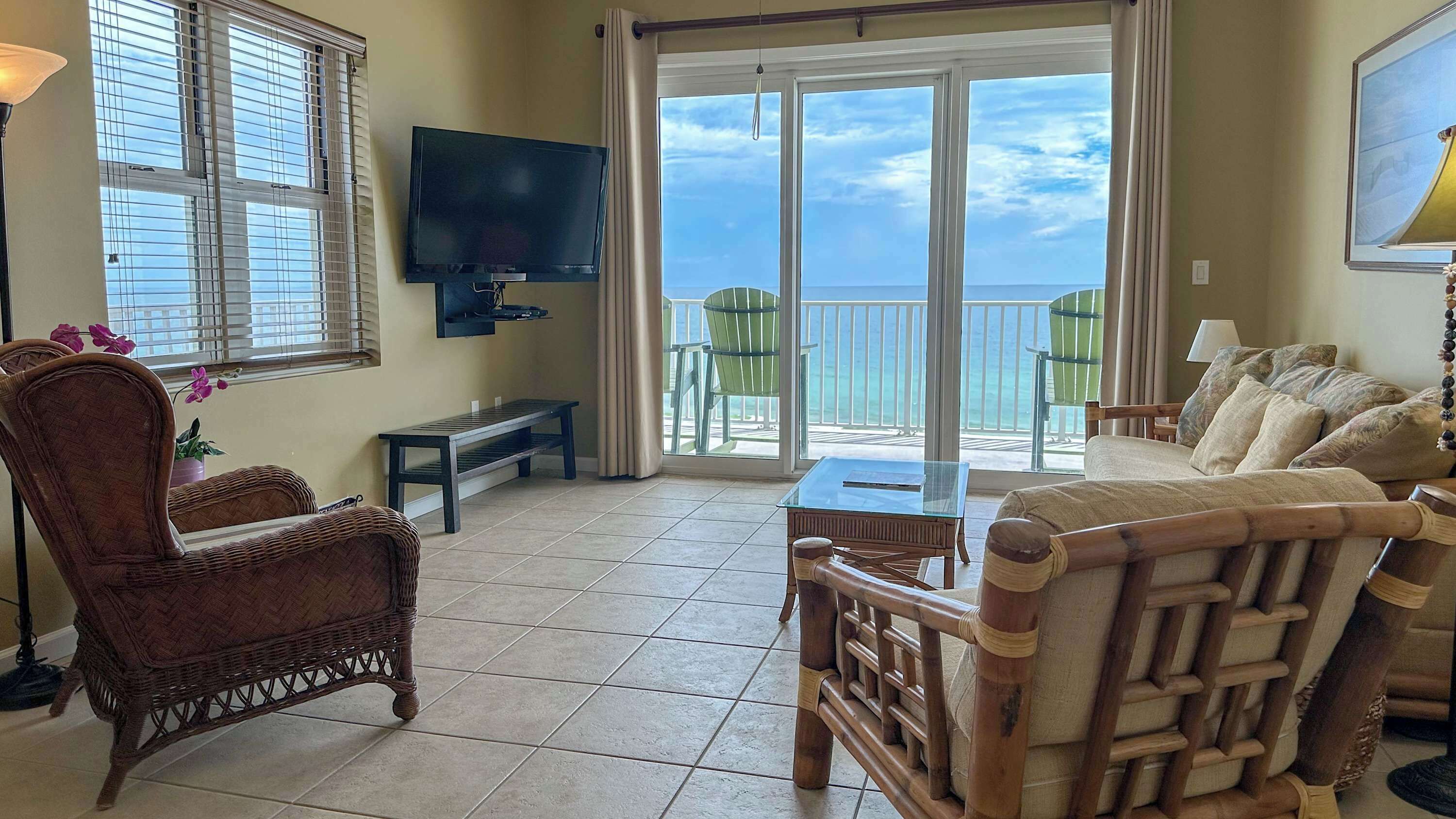 A living room with a large window overlooking the beach, a flat-screen TV mounted on the wall, wicker furniture, and a glass table.