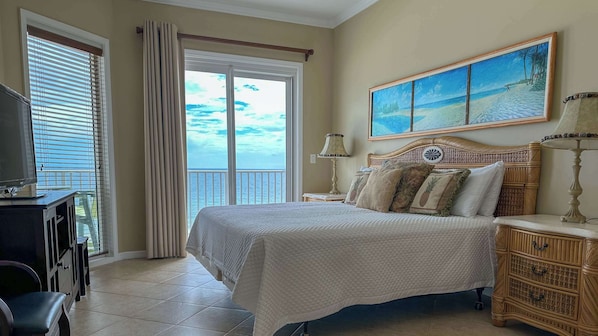 A bedroom with a large bed, a television, a desk, and a view of the ocean through the window.