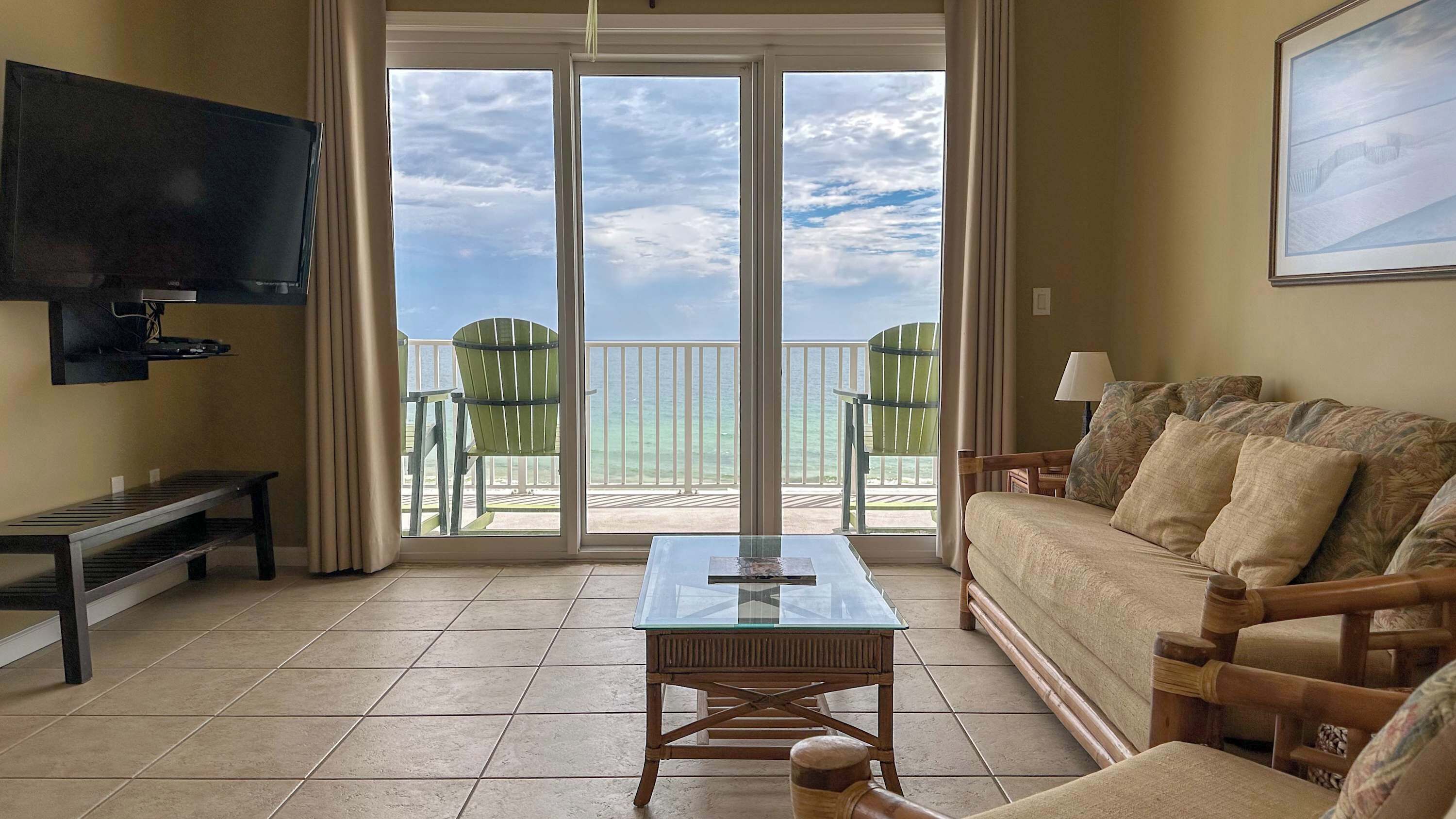 A living room with a TV, a glass-top coffee table, a sofa, and a view of the ocean through large sliding glass doors.