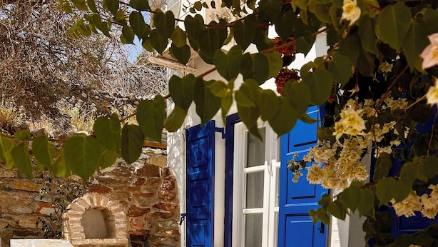 Ein Sitzbereich im Freien aus Stein mit einem Tisch, einem Korbstuhl, blauen Fensterläden und einer Steinmauer.