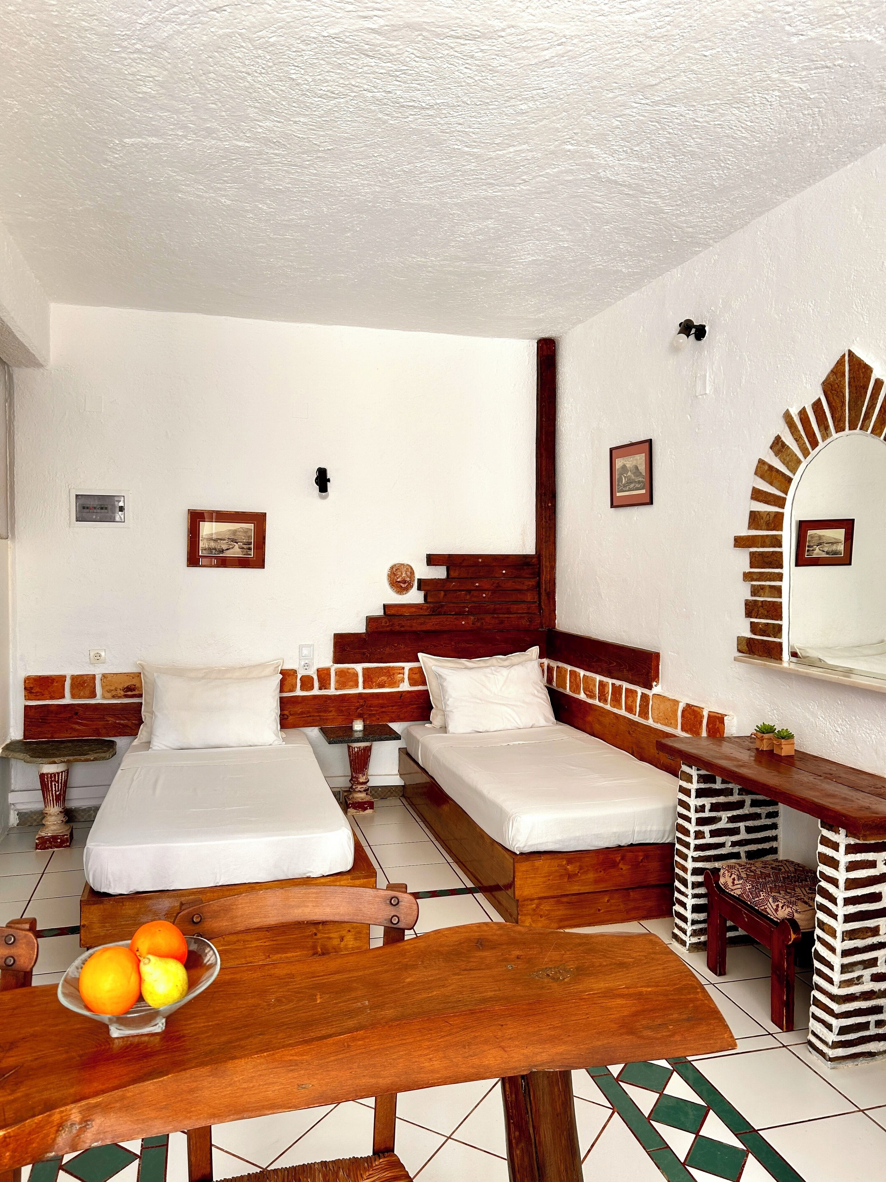 Una habitación con dos camas, un banco de madera, una mesa con fruta y un espejo con marco decorativo.