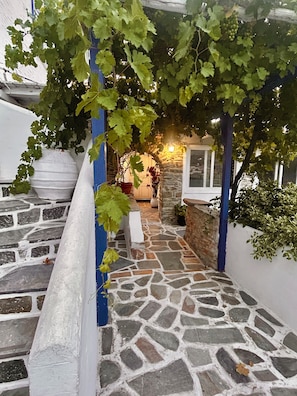 A stone staircase leading to a stone building with a covered area supported by blue columns, a potted plant, and a lit lamp. - Jasmine of Cyclades (Syros)