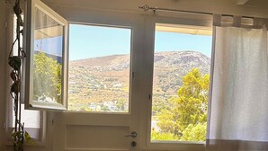 Ein Zimmer mit Blick auf eine hügelige Landschaft durch ein Fenster, eine Zimmerpflanze, eine Steinsitzbank und eine Obstschale auf einem Tisch.
