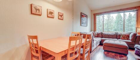 A dining area with a wooden table and chairs, a sofa, and a large window with a view of trees.