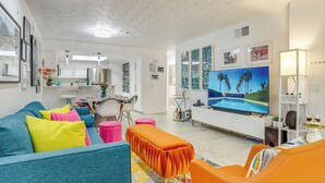 Un salon moderne avec un canapé, un pouf et des fauteuils. On y trouve également un téléviseur et une étagère ornée d’objets décoratifs.