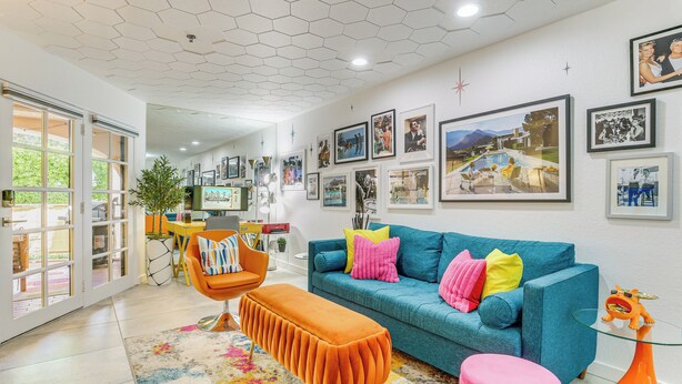 Un salon moderne avec un canapé bleu, un pouf orange et un tabouret rose. Des tableaux encadrés ornent le mur, et un téléviseur est visible au fond.