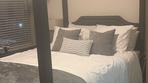 Ein Bett mit Baldachin, ausgestattet mit weißer Bettwäsche und grauen Kissen, ein grauer Teppich und ein Fenster mit Jalousien.