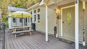 A wooden deck with picnic tables and a yellow umbrella, a door with a decorative sun, and a mailbox. - Riverfront Home w/ Hot Tub Near Killington Resort (Bridgewater Corners)