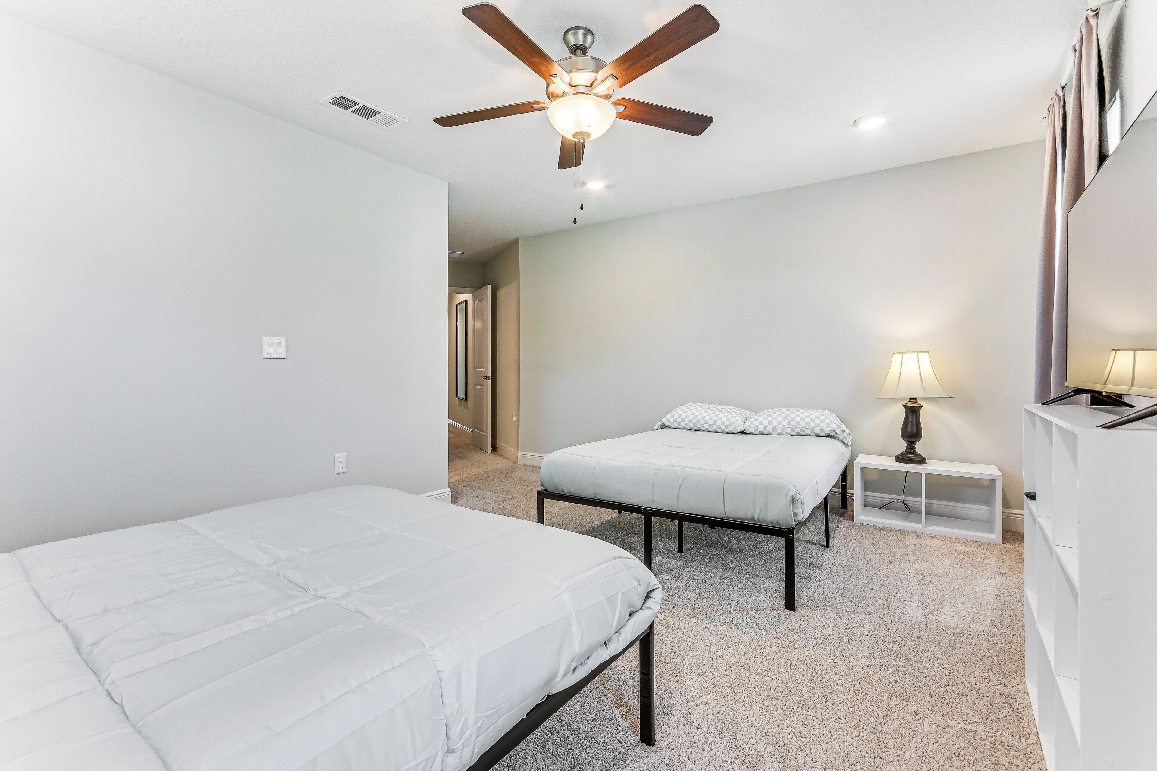 A room with two beds, a ceiling fan, a nightstand with a lamp, and a white wardrobe.
