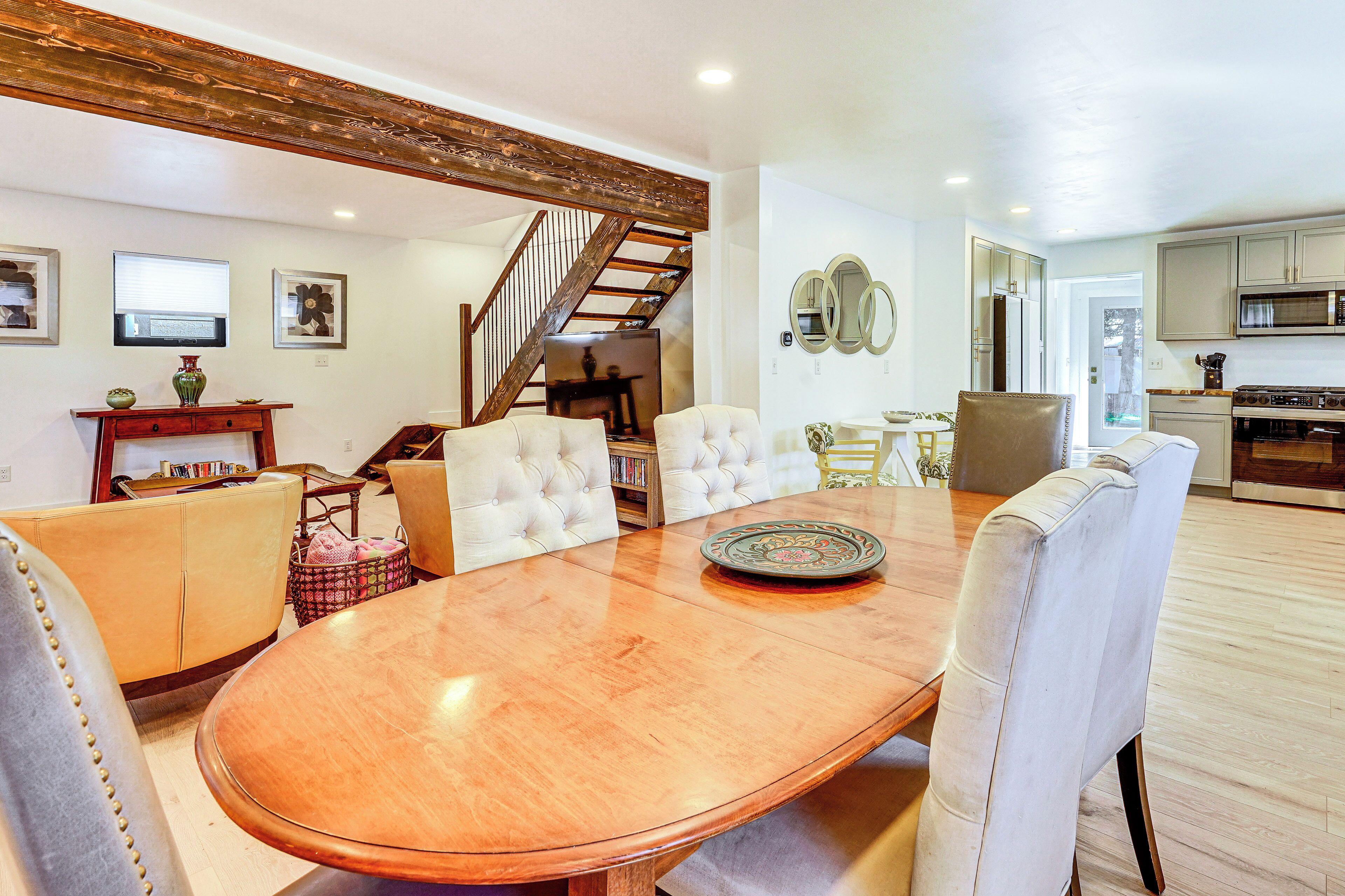 A dining area with a wooden table, white upholstered chairs, a sofa, and a staircase leading to another level.