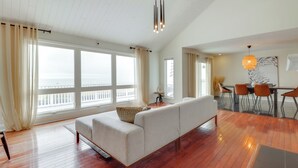 Un vaste espace de vie comprenant un canapé, une table à manger et des chaises, de grandes fenêtres avec des rideaux et une vue sur la mer.
